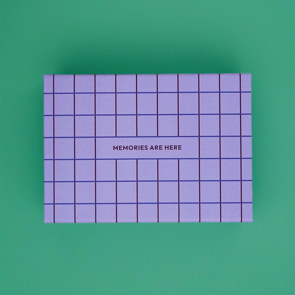Erinnerungskiste – Erinnerungsbox für Babys im Karomuster - Lila von Endless Hugs mit der Aufschrift „MEMORIES ARE Here“ auf dem Deckel ist das ideale Geschenk für Eltern, um Erinnerungen aus dem ersten Lebensjahr zu bewahren. Gezeigt auf grünem Hintergrund.