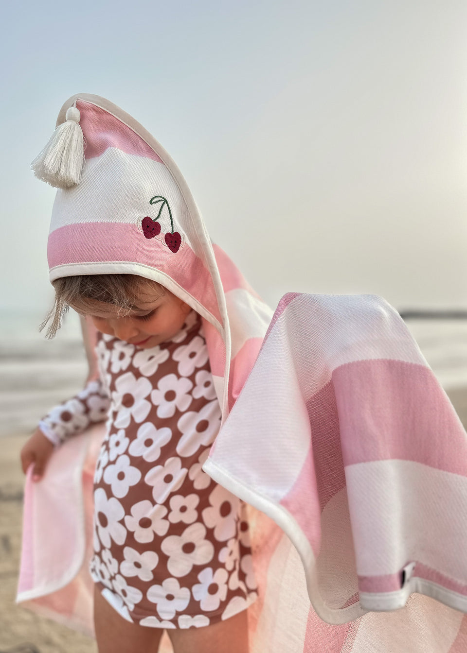 Dziecko w kwiatowym kostiumie kąpielowym stoi na plaży, owinięte w Endless Hugs RĘCZNIK DLA NIEMOWLAKA Z KAPTURKIEM - BAMBUSOWY W PASY / WISIENKI, miękki ręcznik z kapturem wykonany z mieszanki bambusa i bawełny w różowe paski.