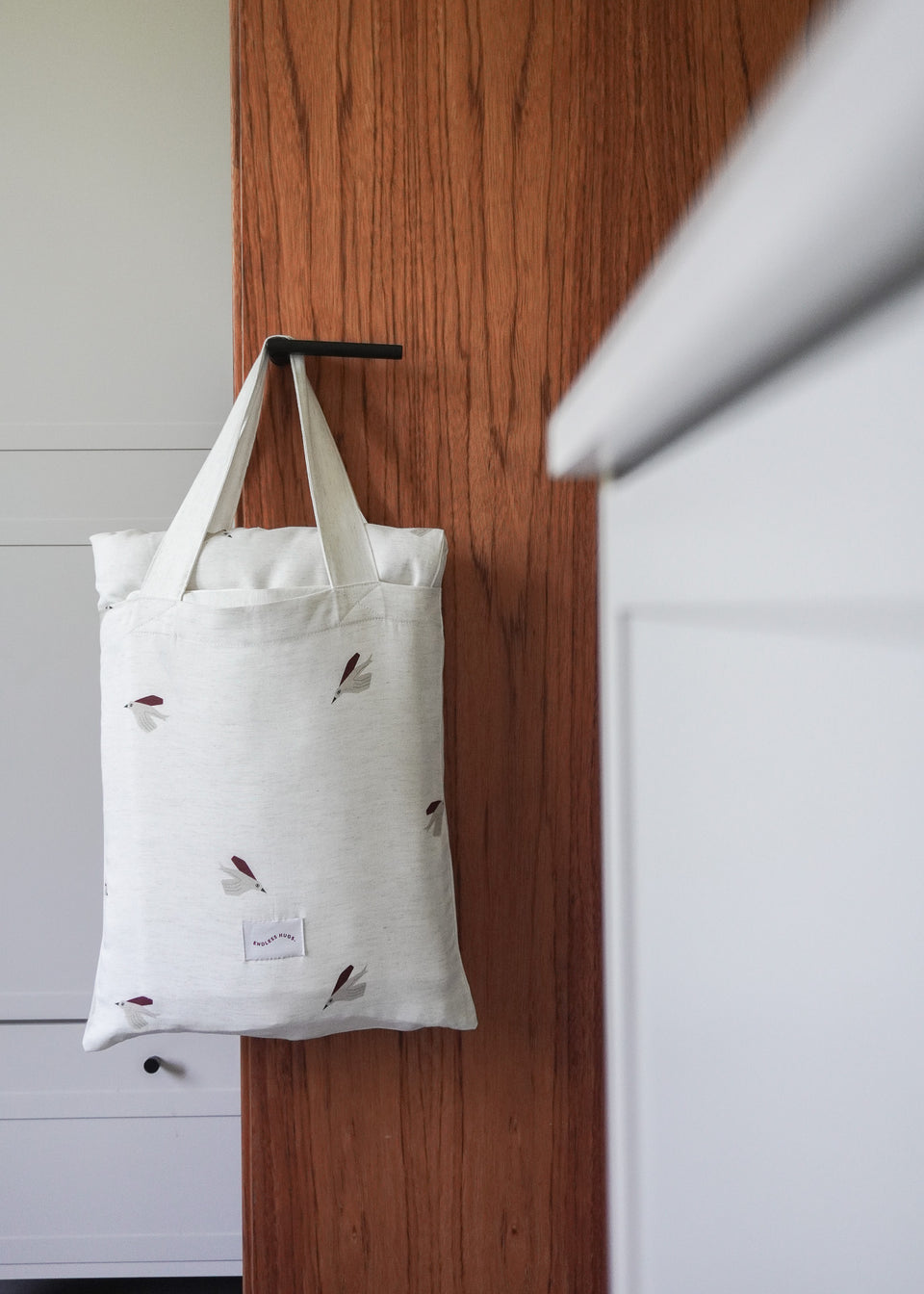 Produkt Endless Hugs, bambusowa torba typu tote POŚCIEL DZIECIĘCA - BAMBUSOWA / PTASZKI w kolorze białym z motywem liści, wisi na czarnym haczyku na drewnianej ścianie.