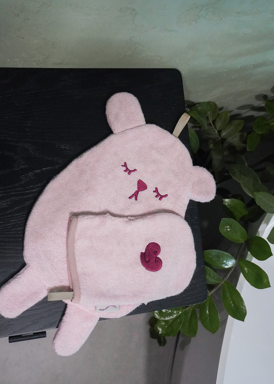 RĘCZNICZEK BAMBUSOWY BEAR, różowy dywanik łazienkowy w kształcie misia i pasująca do niego myjka marki Endless Hugs, wykonane z frotte bambusowej, przedstawione są na ciemnej powierzchni z zielonymi liśćmi.