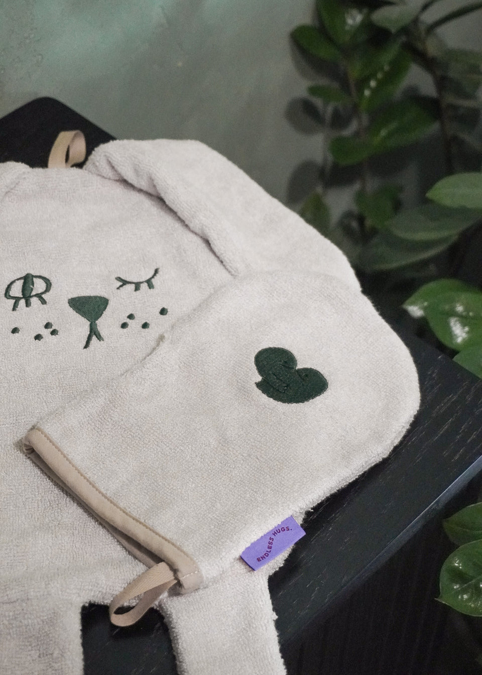 Ręcznik Endless Hugs MYJKA DLA NOWORODKA - BAMBUSOWA - BEŻOWA z twarzą zwierzęcia i pasująca do niego antyalergiczna myjka, ozdobiona motywem serca, umieszczone na ciemnej drewnianej powierzchni otoczonej jaskrawozielonymi liśćmi.