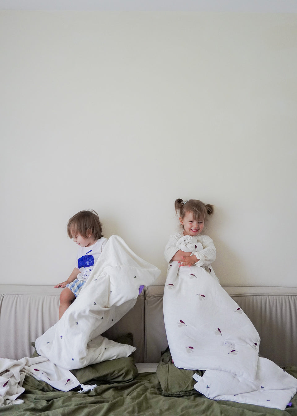 Dwoje małych dzieci siedzi na łóżku, otulonych miękkim materiałem Endless Hugs POŚCIEL DZIECIĘCA - BAMBUSOWA / PTASZKI. Kojący uścisk antyalergicznych włókien bambusowych zapewnia delikatny dotyk na ich skórze, gdy opierają się o gładką ścianę.