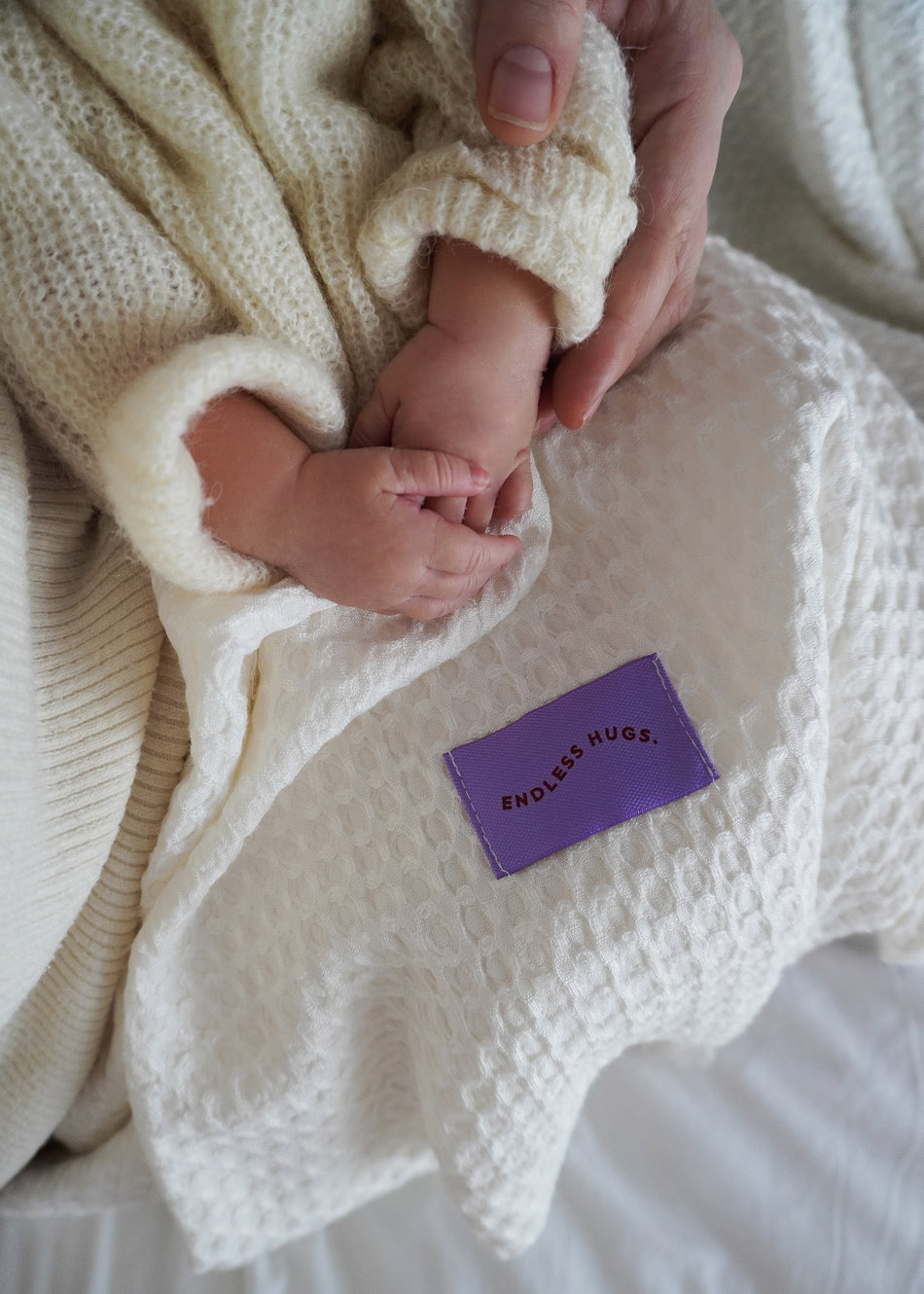 Dłoń dziecka jest delikatnie trzymana przez osobę dorosłą na miękkim, kremowym kocyku bambusowym z chwostami. Kocyk ma fioletową łatkę z napisem Endless Hugs, a obie ręce spoczywają na teksturowanej tkaninie.