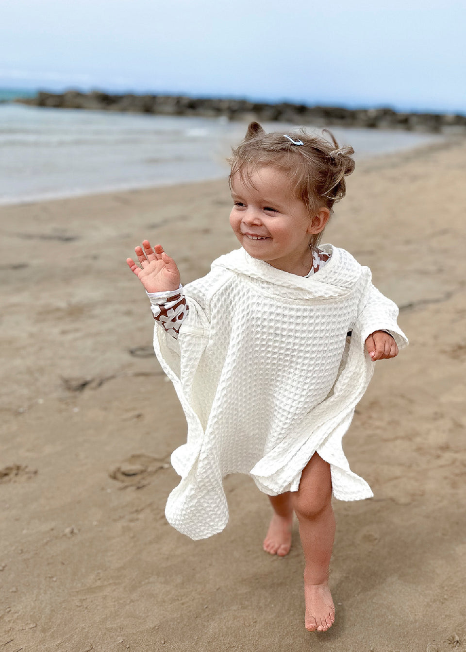Maluch w PONCZO BAMBUSOWYM / PTASZEK marki Endless Hugs biegnie boso po piaszczystej plaży, uśmiechnięty, na tle oceanu i skał.