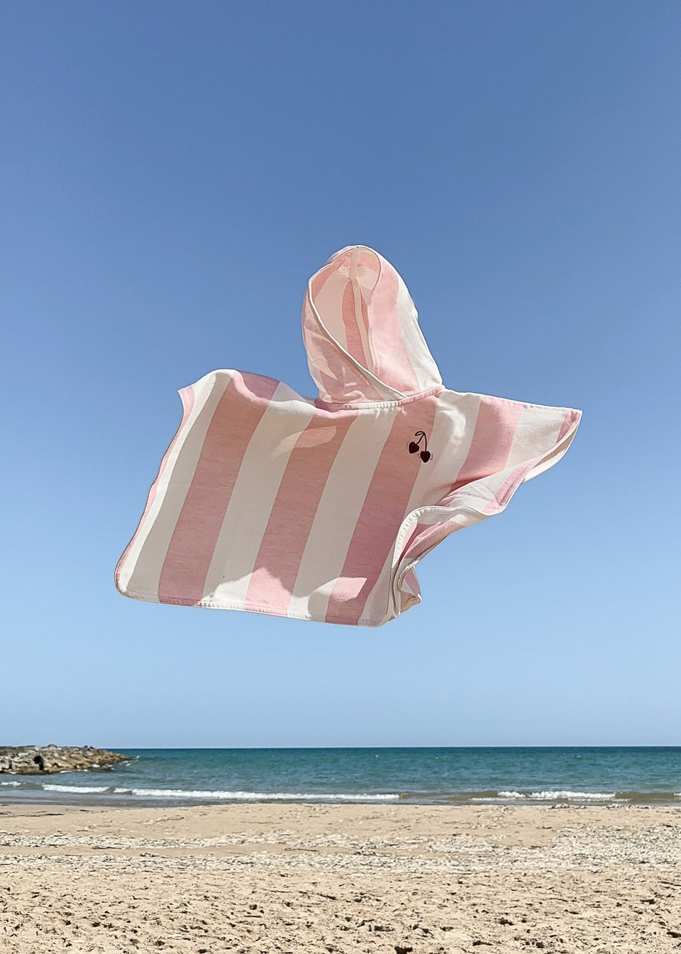 PONCHO AUS BAMBUS IN ROSA STREIFEN / KIRSCHEN der Marke Endless Hugs, aus Bambusviskose gefertigt, schweben am Strand in der Luft vor einem klaren, blauen Himmel.