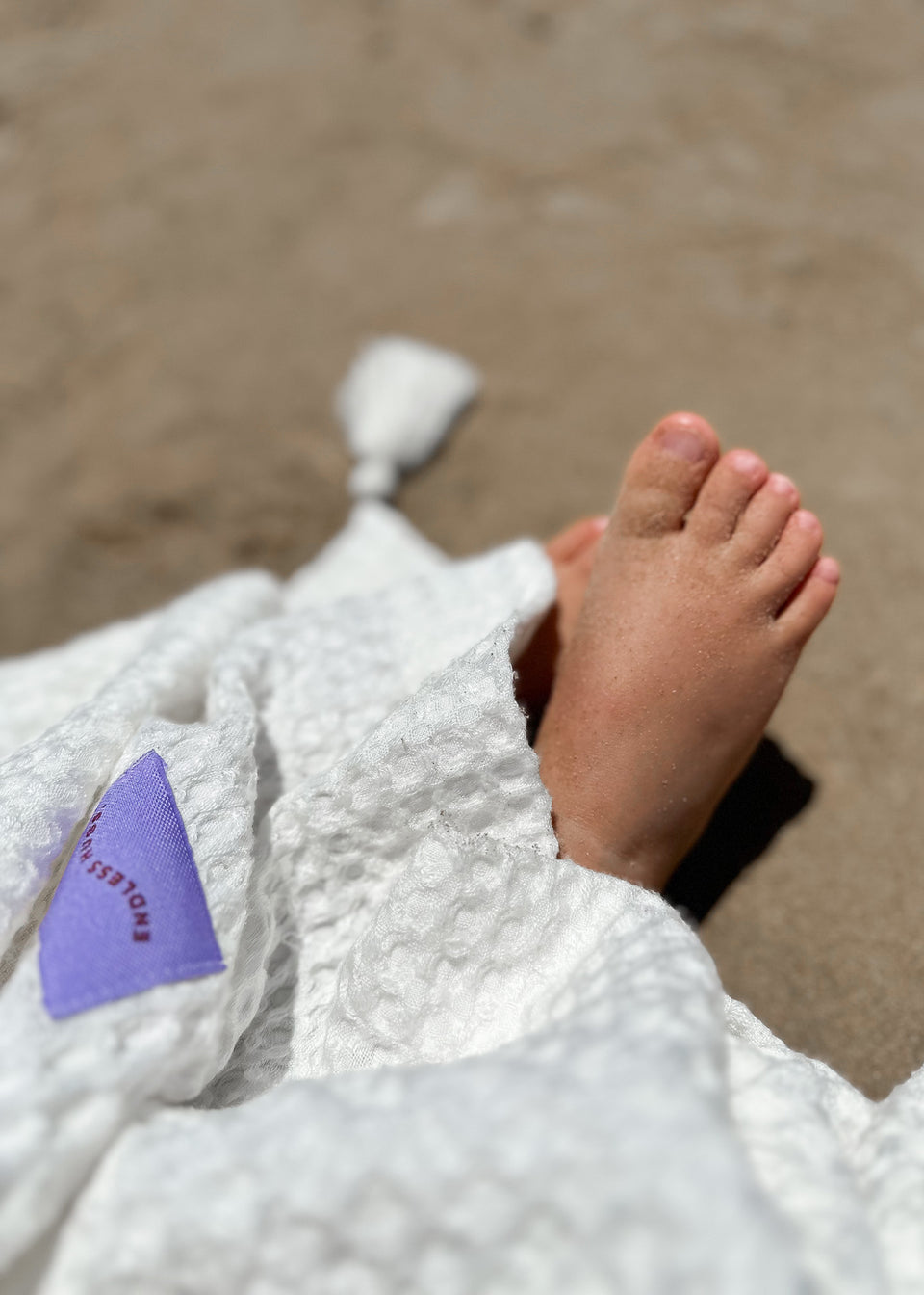 Zbliżenie pokazuje piaszczystą stopę częściowo owiniętą w biały, teksturowany Kocyk bambusowy z chwostami - 100x120 CM na plaży. Na kocu widoczna jest fioletowa metka.