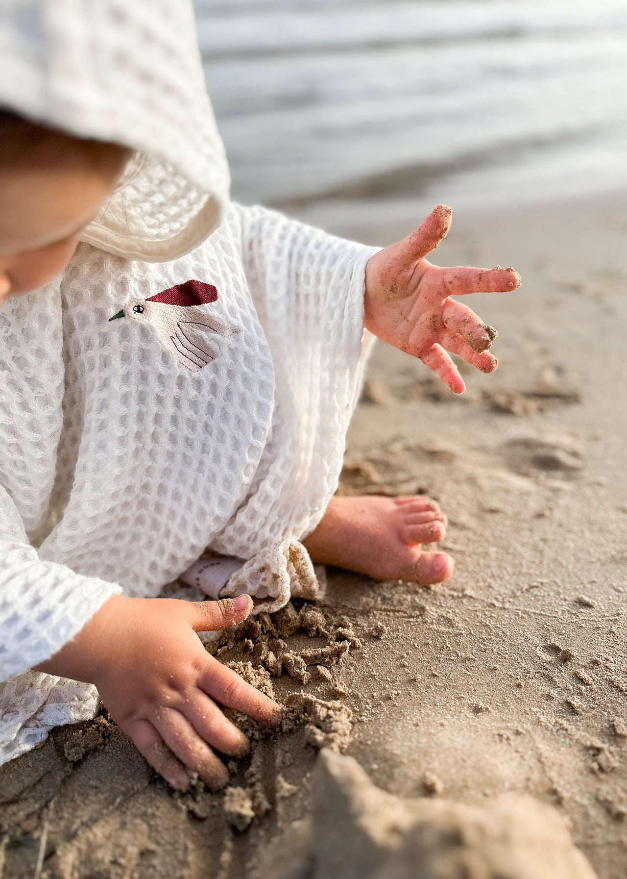Dziecko w białym PONCZO BAMBUSOWYM / PTASZEK Endless Hugs radośnie bawi się piaskiem na plaży, oddając się beztroskiemu duchowi lata.