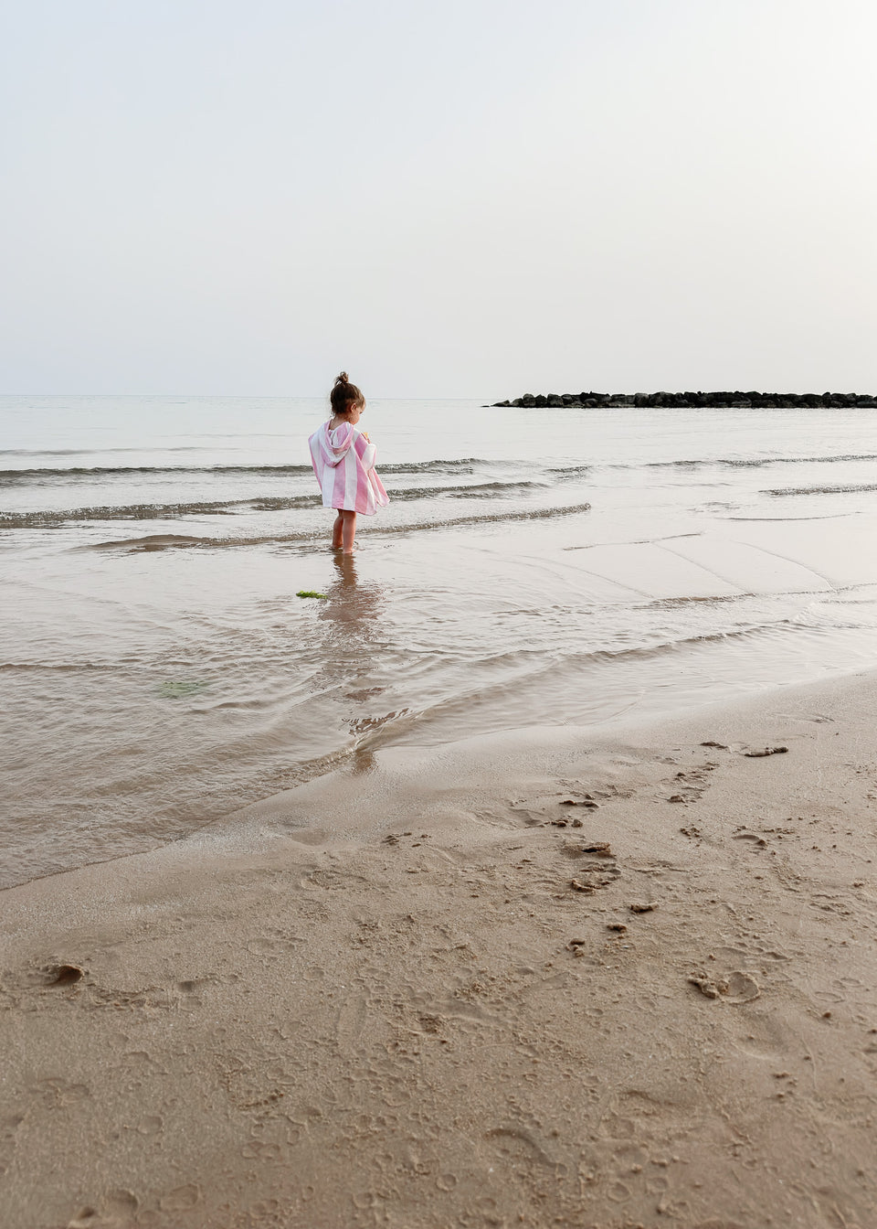 Das Kind steht am Wasserufer am Strand, eingewickelt in einen BAMBUS-PONCHO MIT ROSA STREIFEN / KIRSCHEN von Endless Hugs, mit dem Gesicht zum Meer unter einem bewölkten Himmel.