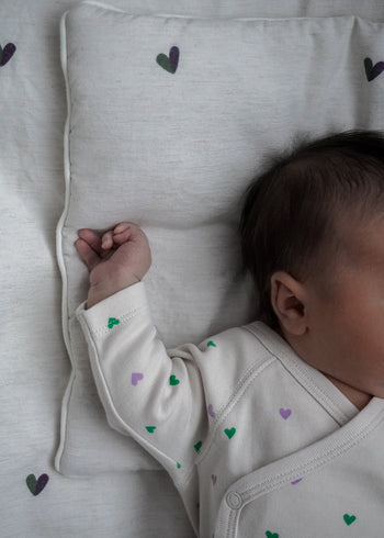 Niemowlę śpi na POŚCIELU DZIECIĘCA - BAMBUSOWA / SERDUSZKA firmy Endless Hugs, ubrane w lekki strój z zielonymi i fioletowymi serduszkami. Pościel bambusowa, znana z hipoalergicznych i termoregulujących tkanin, wzmacnia spokojny sen.