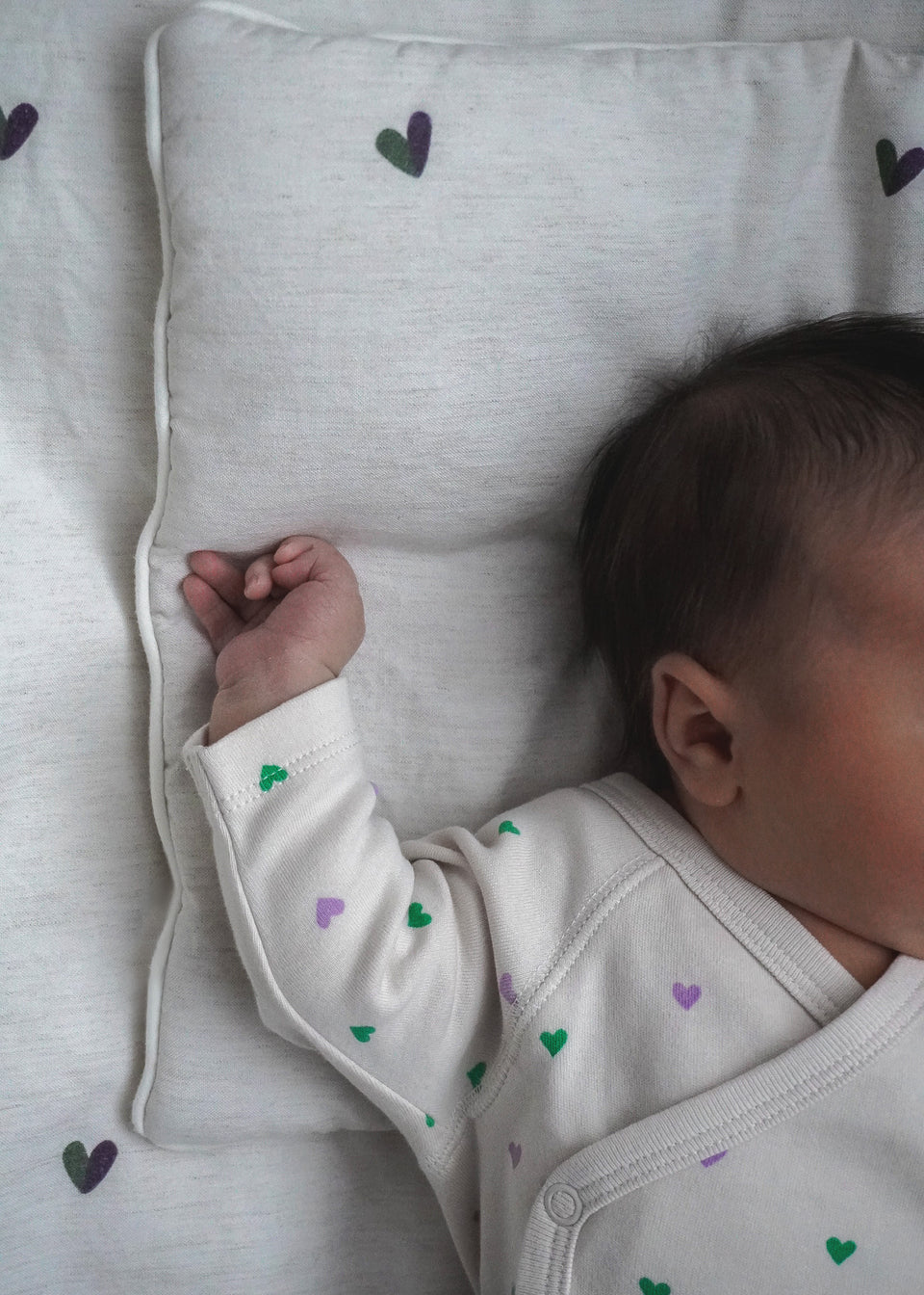 Zbliżenie na niemowlę na Endless Hugs PODUSZKA BAMBUSOWA / SERDUSZKA, ubrane w jednoczęściowy kombinezon z zielonymi i fioletowymi serduszkami. Twarz niemowlęcia jest częściowo widoczna, jedno ramię uniesione, cieszące się hipoalergicznymi zaletami bambusa.