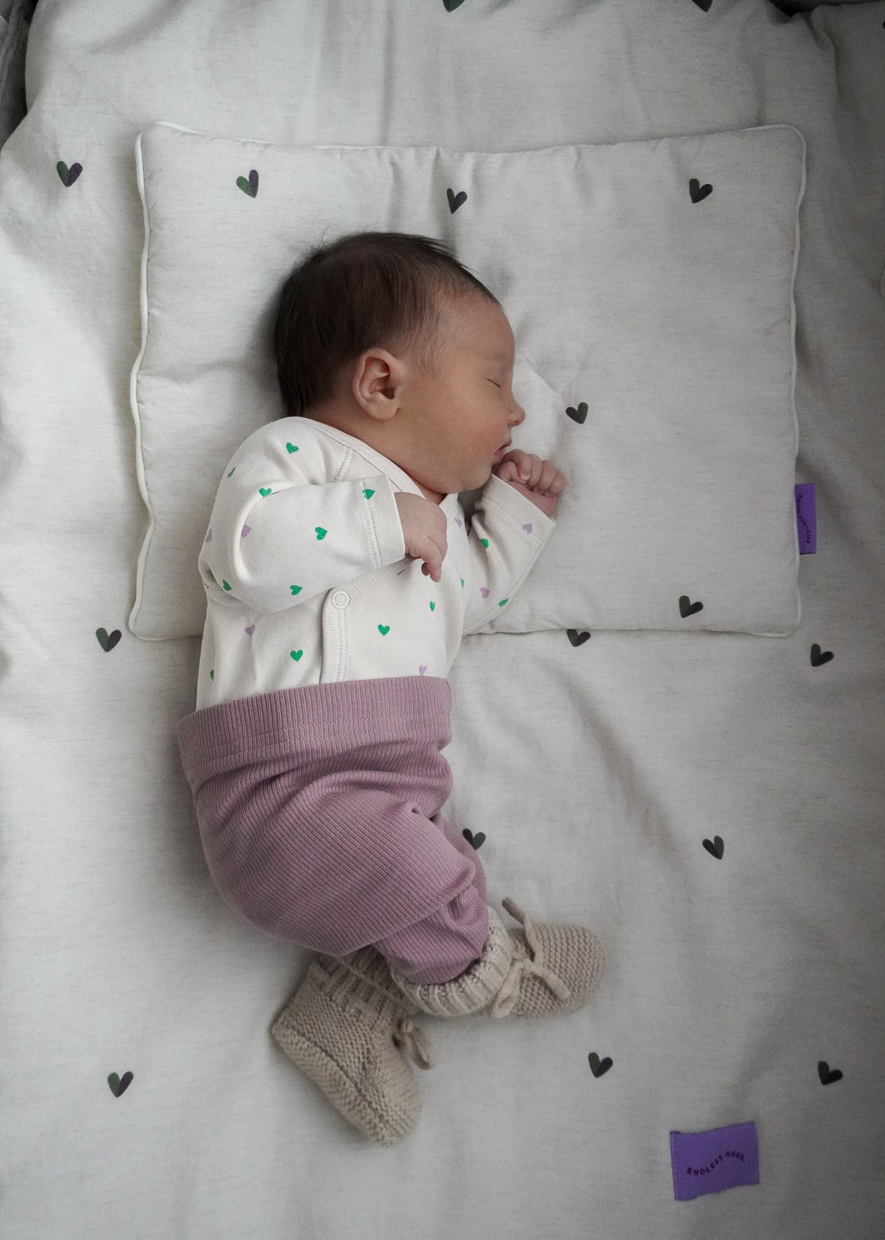 Niemowlę śpi na poduszce i kocyku POŚCIEL DZIECIĘCA - BAMBUSOWA / SERDUSZKA firmy Endless Hugs, ubrane w biały pajacyk w zielone kropki, liliowe spodnie i dzianinowe buciki.