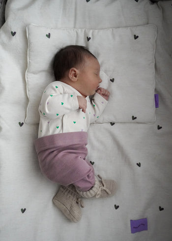 Niemowlę śpi na poduszce i kocyku POŚCIEL DZIECIĘCA - BAMBUSOWA / SERDUSZKA firmy Endless Hugs, ubrane w biały pajacyk w zielone kropki, liliowe spodnie i dzianinowe buciki.