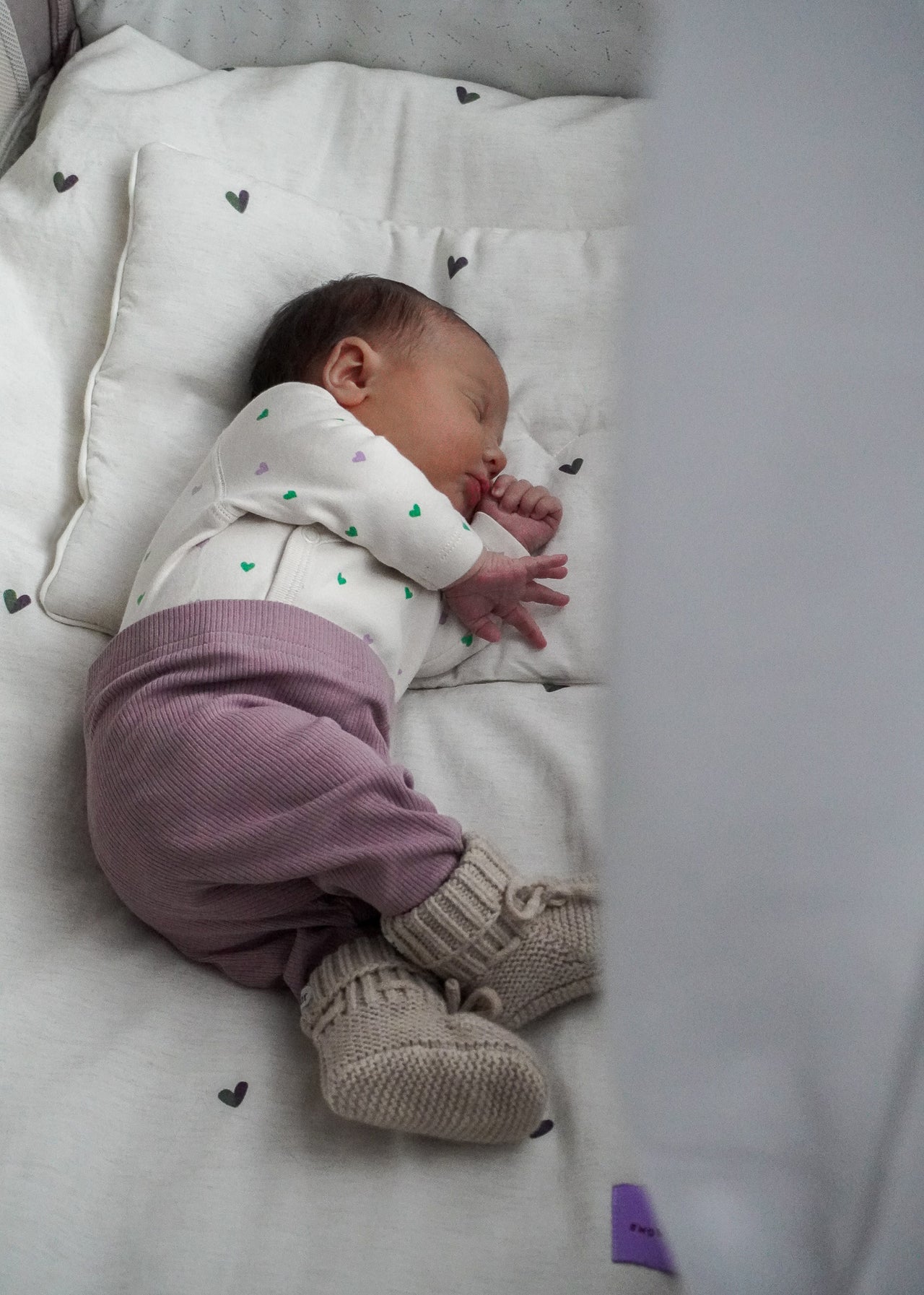 Niemowlę śpi spokojnie na białej poduszce, ubrane w białą koszulę z serduszkami, fioletowe spodnie i beżowe dzianinowe buciki. Otulone hipoalergiczną pościelą bambusową Endless Hugs POŚCIEL DZIECIĘCA - BAMBUSOWA / SERDUSZKA dla dzieci.