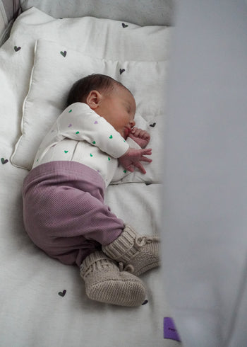 Niemowlę śpi spokojnie na białej poduszce, ubrane w białą koszulę z serduszkami, fioletowe spodnie i beżowe dzianinowe buciki. Otulone hipoalergiczną pościelą bambusową Endless Hugs POŚCIEL DZIECIĘCA - BAMBUSOWA / SERDUSZKA dla dzieci.