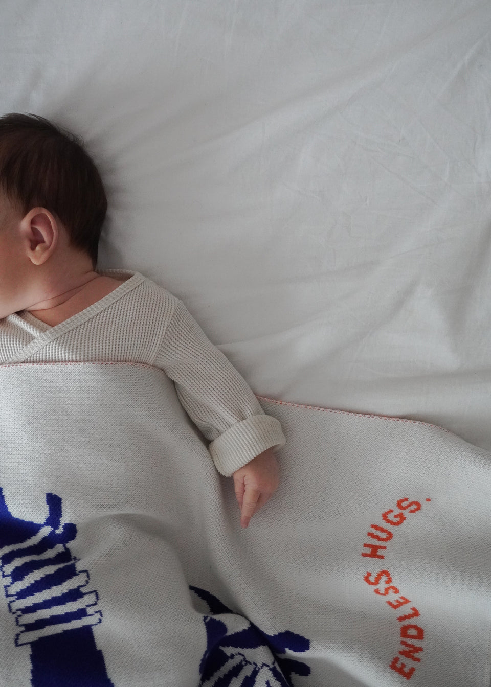Spokojnie śpiące dziecko jest częściowo przykryte bawełnianym kocykiem dziecięcym „Endless Hugs” z uroczym niebieskim wzorem z pieskiem.