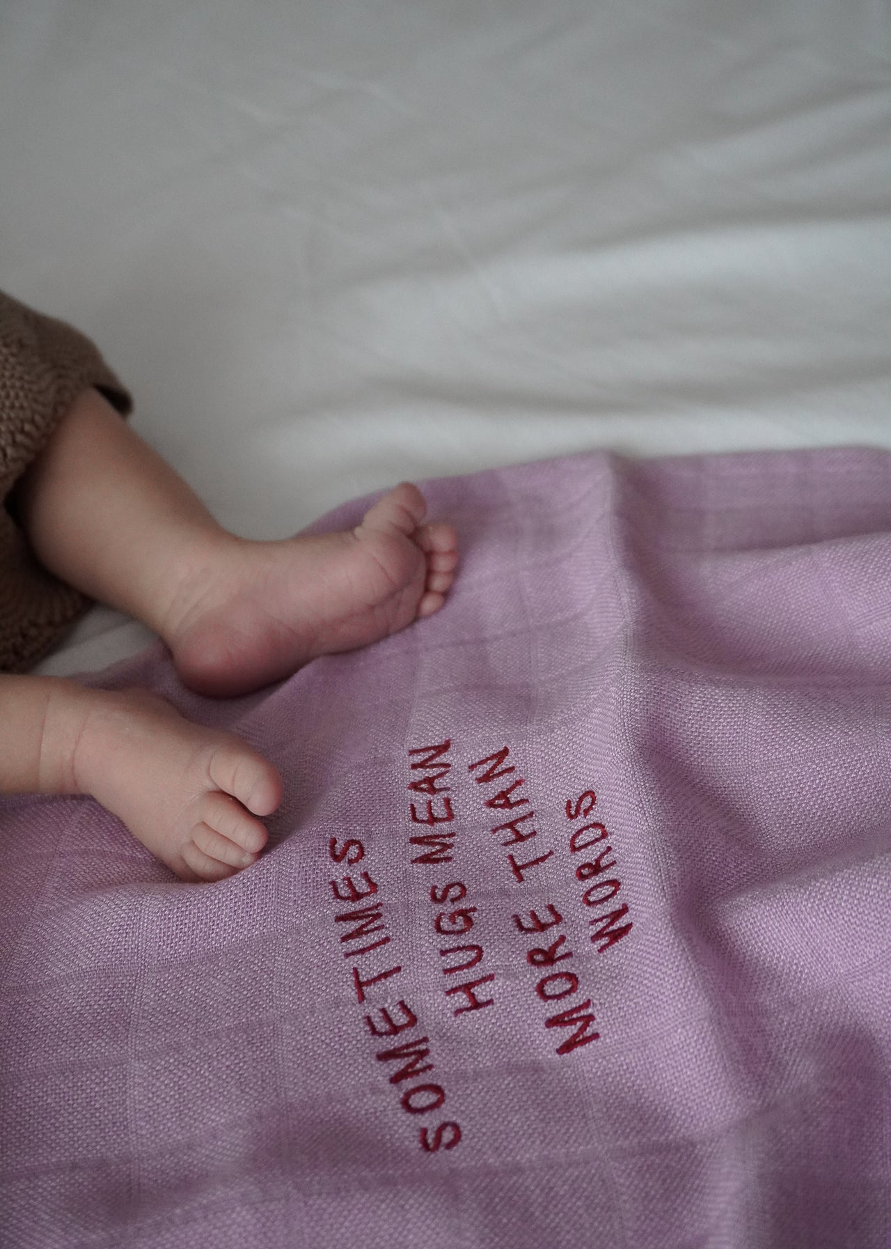 Stopy dziecka na różowym bambusowym OTULACZU DLA NOWORODKA od Endless Hugs z haftowanym napisem „CZASAMI PRZYTULASY OZNACZAJĄ WIĘCEJ NIŻ SŁOWA”. Ten niezbędny element o wymiarach 120x120 cm w kolorze fioletowym zapewnia właściwości termoregulacyjne, aby otulić noworodka komfortem i miłością.