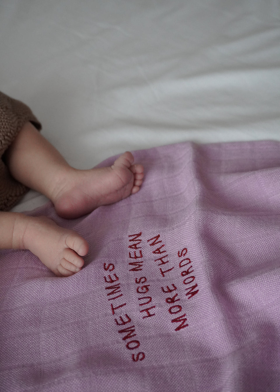 Stopy dziecka na różowym bambusowym OTULACZU DLA NOWORODKA od Endless Hugs z haftowanym napisem „CZASAMI PRZYTULASY OZNACZAJĄ WIĘCEJ NIŻ SŁOWA”. Ten niezbędny element o wymiarach 120x120 cm w kolorze fioletowym zapewnia właściwości termoregulacyjne, aby otulić noworodka komfortem i miłością.